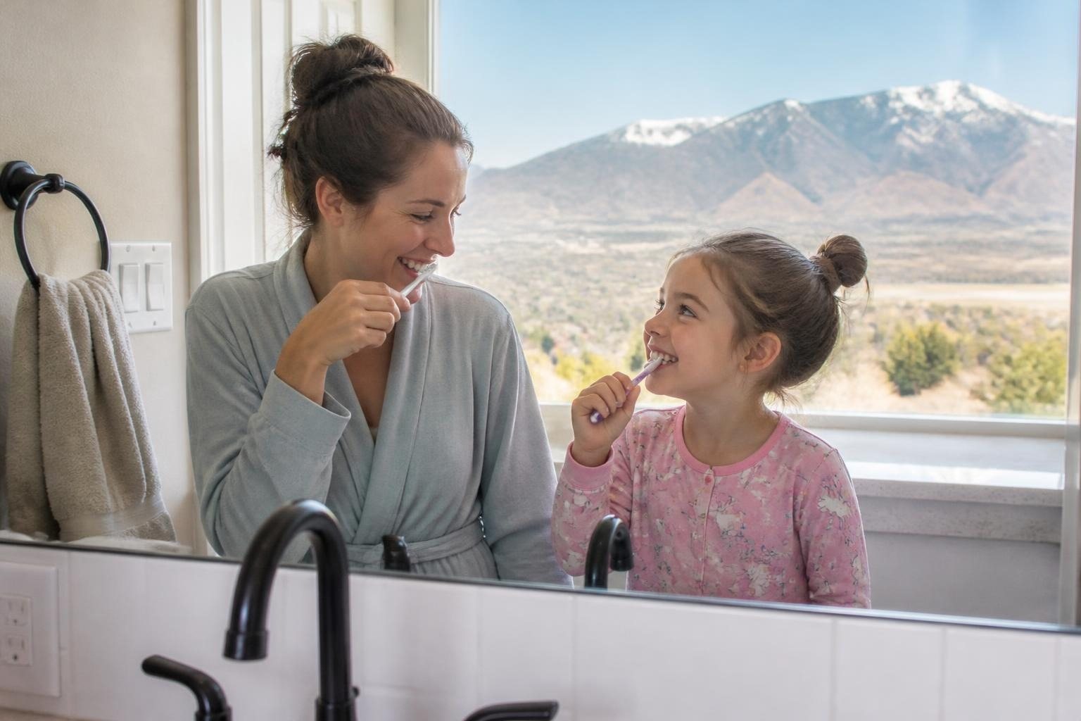 A mother and young daughter brushing their teeth together, with Mount Nebo outside the window